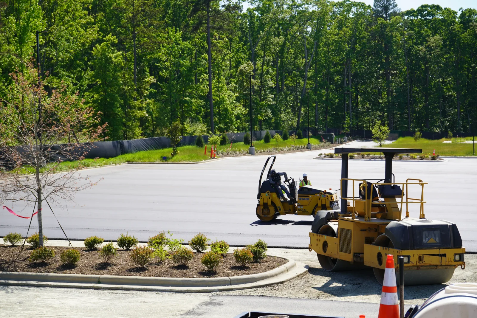 Asphalt paving equipment compacting fresh asphalt on commercial parking lot with concrete curb and landscaping