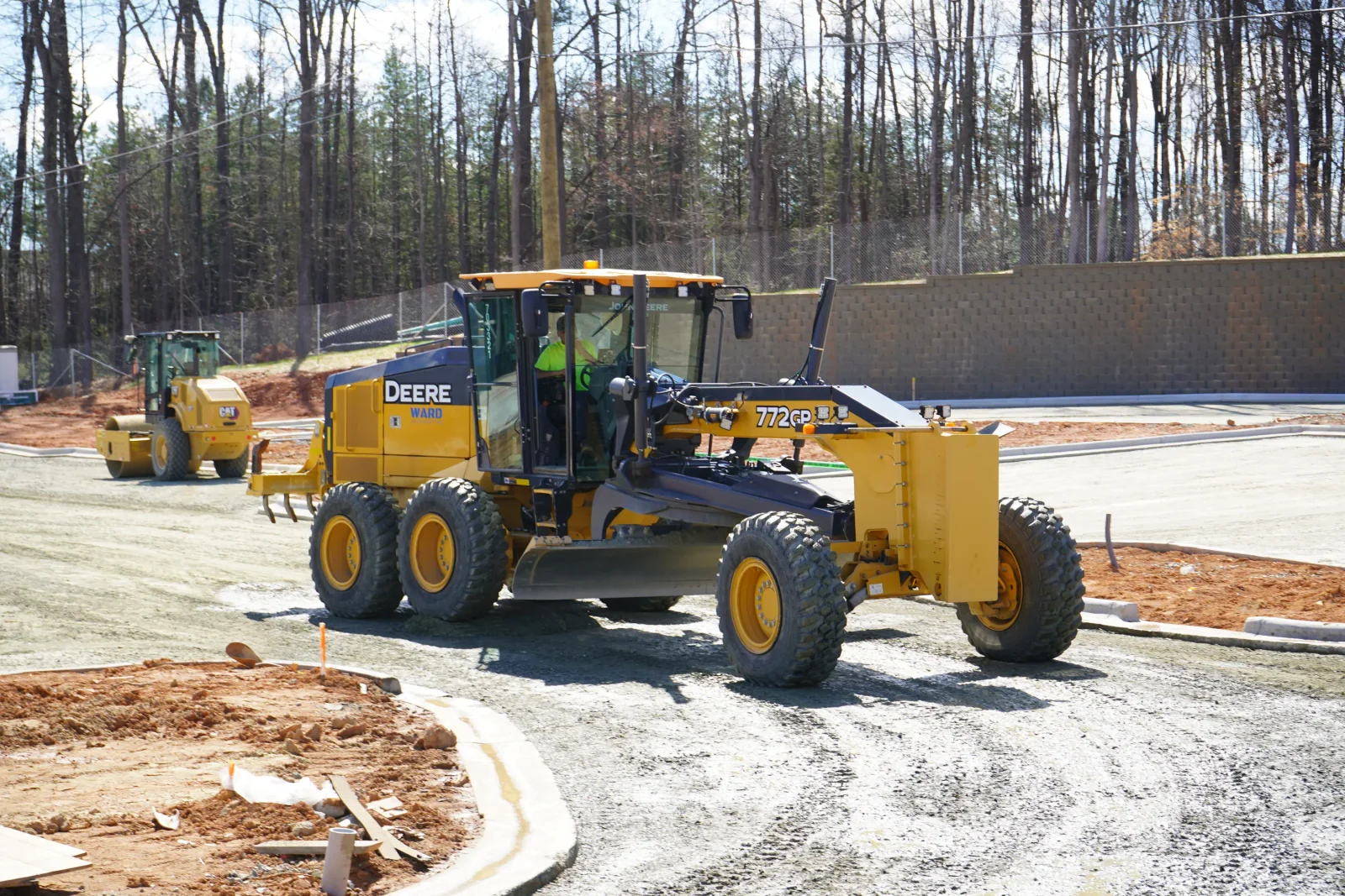Motor grader and compaction equipment grading dirt site with concrete curbing and retaining wall